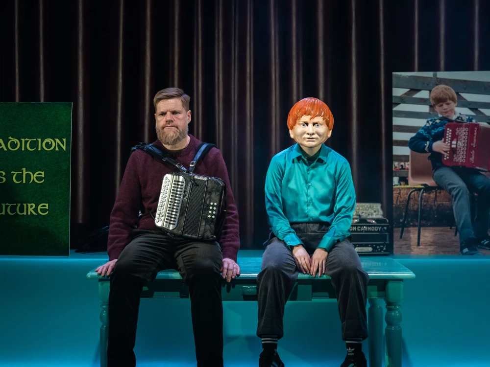 Photo of two actors sitting on a table - one masked.