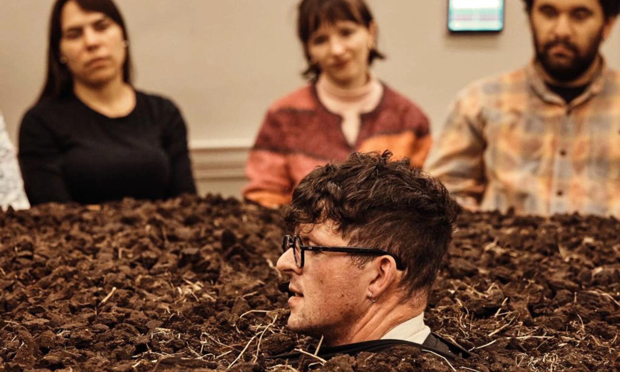 Photo of man's head emerging from peat-covered table with people around at theatre performance