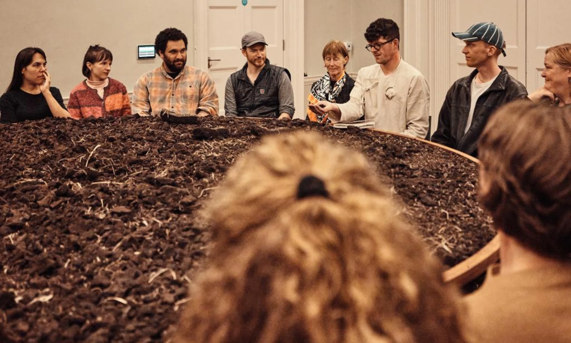 Photo of man's head emerging from peat-covered table with people around at theatre performance