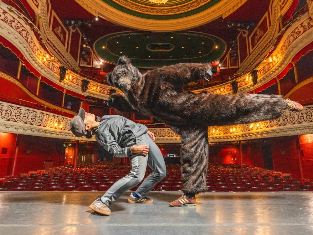 Photo of two actors on theatre set dancing, one in a bear costume.