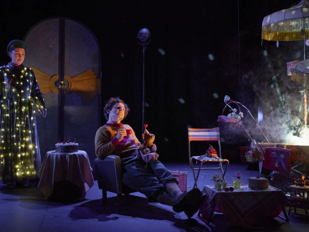 Stage scene with man sitting and female standing on left.