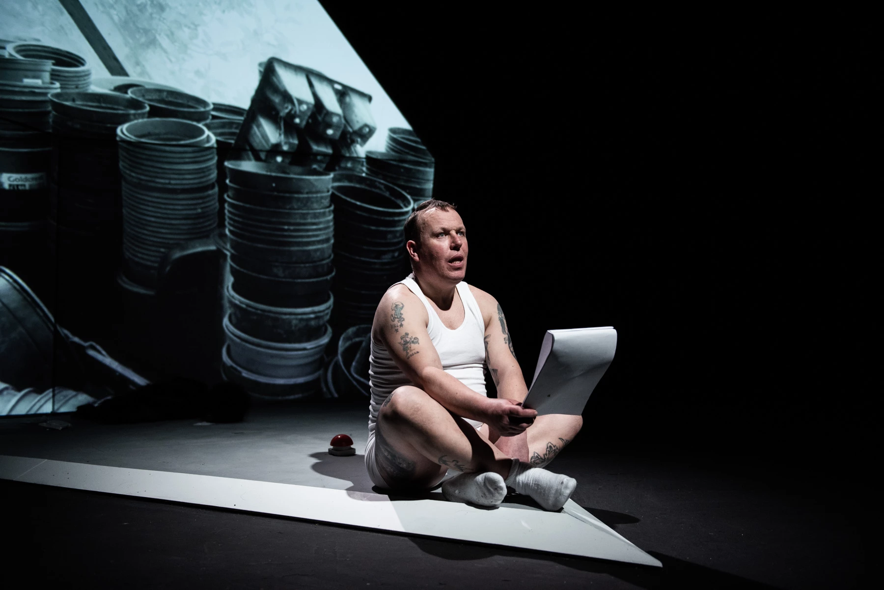 Man sitting cross-legged on stage with a projection of plant pots behind him. 