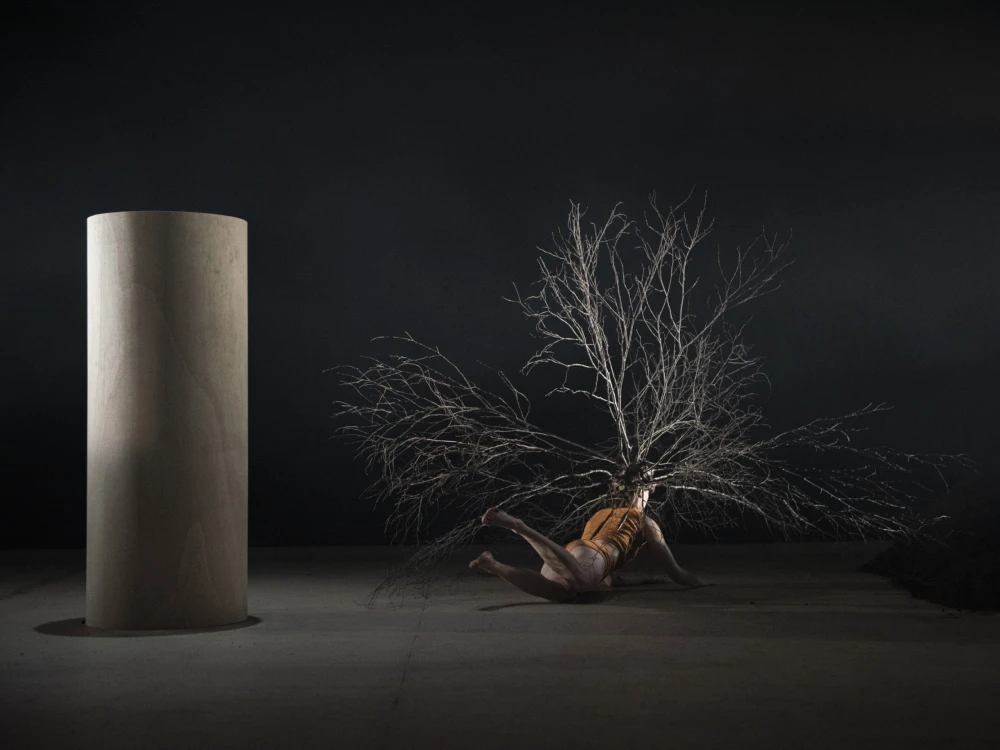 Stage with large pillar on left and woman with large headpiece on ground on right.