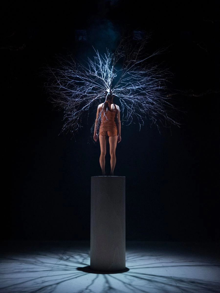 Woman with large headpiece standing on large pillar on a dark stage.